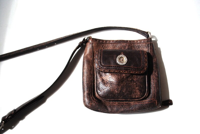 May include: A brown leather crossbody bag with a flap closure and a silver button. The bag has a front pocket with a decorative stitch detail.