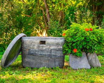 May include: A gray stone fire pit with a lid, flames burning inside, and a small bush with red flowers in the background.