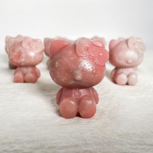 Puede incluir: Varias figuras de piedra rosa con forma de gato con lazo. Las figuras son de color rosa claro con un aspecto ligeramente translúcido. Las figuras de gato están de pie con los brazos a los lados.