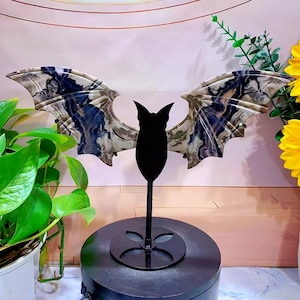 Puede incluir: Una escultura decorativa de murciélago con las alas extendidas. Las alas presentan un patrón veteado en tonos azules, crema y gris. El cuerpo del murciélago es una silueta negra sólida, montada sobre un soporte de metal negro.