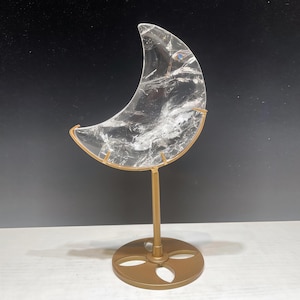 Pode incluir: Uma escultura de lua crescente em quartzo transparente sobre um suporte de metal dourado. A lua tem veios internos e é montada em uma haste fina com uma base circular. A escultura é exibida em um fundo escuro.