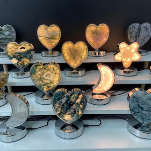 Könnte beinhalten: Eine Sammlung von beleuchteten Lampen in Herz-, Stern- und Halbmondform. Die Lampen zeigen Muster aus Naturstein in Grün, Gold und Weiß. Jede Lampe steht auf einem silberfarbenen Sockel und strahlt ein warmes Licht aus.
