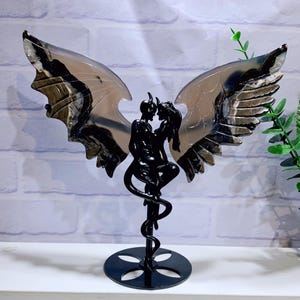 May include: A black metal sculpture of a couple embracing, with devil horns and a tail, and large, patterned wings. The wings are made of a translucent material with black and gray veining. The sculpture is mounted on a black base.