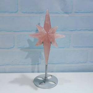 May include: A pink star-shaped crystal ornament on a silver metal stand. The star has six points and a pointed tip. The stand has a circular base with a decorative design. The background is a light blue brick wall.
