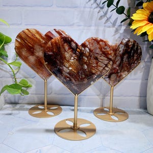 May include: Three heart-shaped decorative objects. Each heart is made of a translucent, brown and white stone with a unique pattern. They are mounted on gold-colored metal stands with circular bases, creating a striking display.