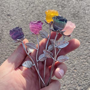 May include: A collection of gemstone rose sculptures in various colors, including purple, pink, yellow, and green. Each rose is mounted on a silver-colored stem with detailed leaf accents. The roses are held in a hand against a blurred background.
