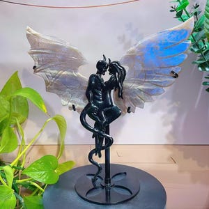Puede incluir: Una escultura negra de una pareja abrazándose, con cuernos y cola, sobre alas iridiscentes. La escultura está montada sobre un soporte negro con una base circular. Las alas tienen un brillo nacarado con reflejos azules.