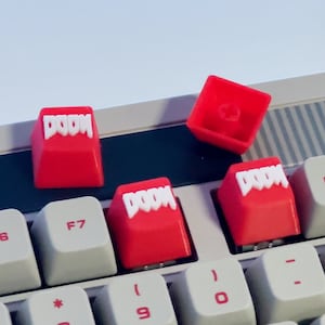 May include: Four red keyboard keycaps with the word "DOOM" printed in white letters. The keycaps are on a white keyboard with black keys.