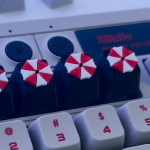 May include: A white mechanical keyboard with black keycaps featuring red and white umbrella symbols. The keyboard has a row of function keys with labels like "SC", "FS", and "SBADO".