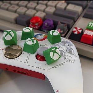 May include: A white and red game controller with green and white Xbox logo keycaps. Other keycaps feature various designs and colors, including red, purple, and blue. A keyboard is in the background.
