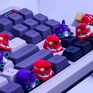 May include: A white mechanical keyboard with purple, red, and green keycaps featuring anime characters. The keycaps are shaped like the heads of robots with red, green, and purple accents. The keyboard has a gray and white color scheme.