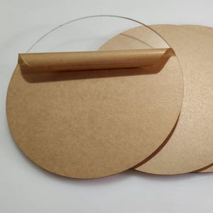 Puede incluir: Una pila de discos redondos de cartón marrón claro con un asa de plástico transparente. Los discos tienen una superficie texturizada y están dispuestos sobre un fondo blanco. El asa está fijada al disco superior.