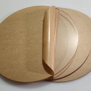 May include: A stack of round, light brown acrylic discs with a protective film partially peeled back. The discs are of varying sizes and appear to be new. The background is a plain white surface.