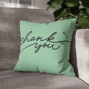 May include: A square, pale green cushion featuring the words "thank you" in black cursive writing. The cushion is placed on a grey sofa. This is a decorative item.