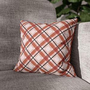 Puede incluir: Un cojín cuadrado decorativo con un diseño de cuadros en tonos naranja, rojo, negro y crema. El cojín está colocado sobre una silla tapizada gris, ofreciendo un acento acogedor y elegante para la decoración del hogar.