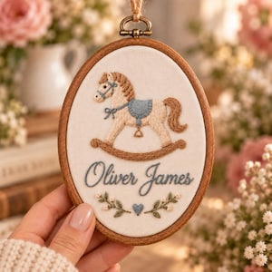 May include: An oval wooden embroidery hoop containing a cream-colored fabric panel. The fabric features a stitched rocking horse in tan and blue, the name Oliver James in blue cursive script, and a small blue heart with floral sprigs.