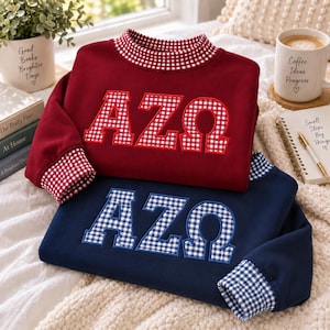 Puede incluir: Dos sudaderas de manga larga con cuello redondo, una en color granate y otra en azul marino. Ambas cuentan con cuello y puños de cuadros vichy rojos y blancos o azules y blancos, y letras griegas AZΩ en tela de cuadros a juego.