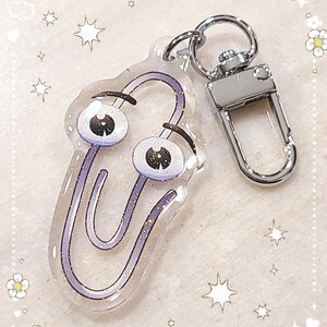 May include: A purple glitter paperclip keychain with googly eyes. The keychain is attached to a silver metal key ring.