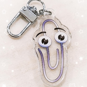 May include: A silver key chain with a clear acrylic charm featuring a purple paperclip with a face. The paperclip has large eyes and a furrowed brow.