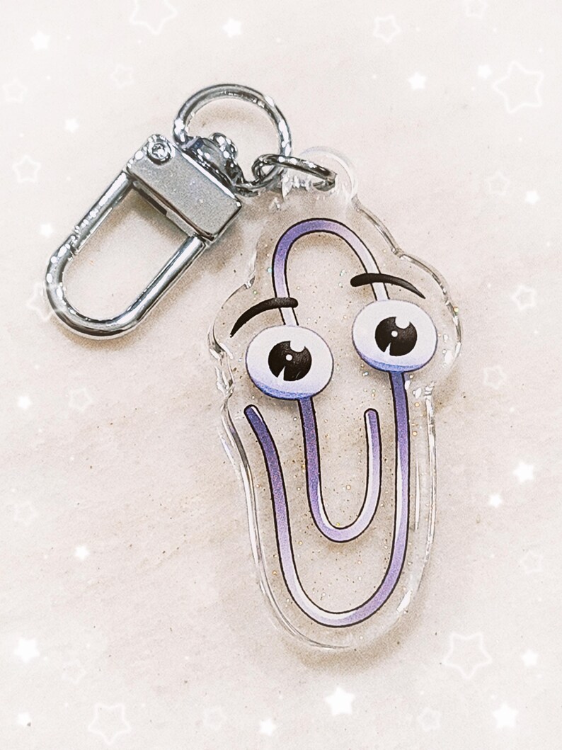 May include: A silver key chain with a clear acrylic charm featuring a purple paperclip with a face. The paperclip has large eyes and a furrowed brow.