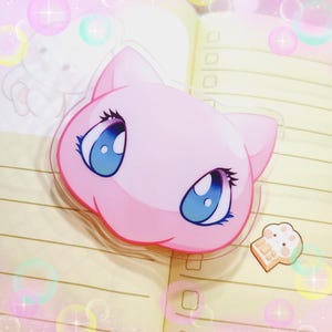 May include: Pink and blue cartoon character with large eyes and a rounded face. The character is a clear acrylic charm.