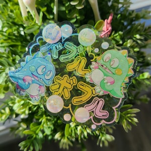 Peut inclure: Un charme transparent et iridescent avec un motif de dessin animé coloré. Le charme présente deux personnages de dessin animé, un dinosaure bleu et vert, et le texte "Bubble Bobble" en japonais.