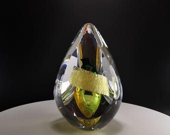 Op de afbeelding: Een transparante glazen sculptuur in de vorm van een traan met bladgoud, groen en geel glas erin.