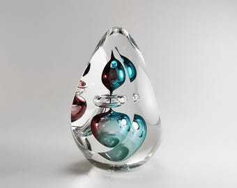 May include: A clear glass paperweight with a teardrop shape. Inside the paperweight are several bubbles of teal and burgundy glass.