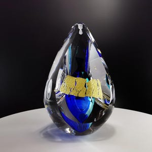 Op de afbeelding: Een heldere glazen vaas met een traanvorm, met blauwe en groene glazen accenten en een band van bladgoud.