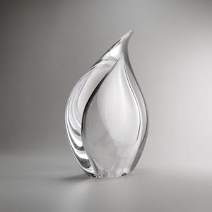 Può includere: Una scultura in vetro trasparente a forma di goccia o di fiamma. L'oggetto presenta superfici lisce e curve che catturano la luce, creando sottili riflessi e ombre. Il vetro è trasparente e solido, con una parte superiore appuntita e base.
