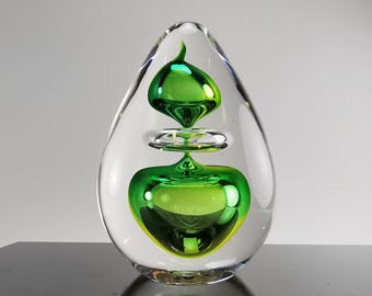 May include: A clear glass sculpture with a teardrop shape. The sculpture contains three green glass orbs, one inside the other. The orbs are iridescent and reflect light.