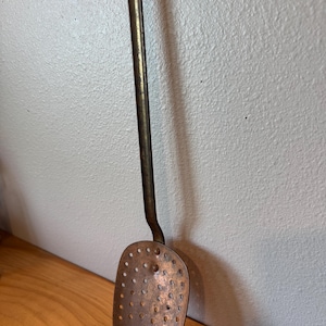May include: A vintage copper skimmer with a long handle and a perforated, oval-shaped head. The handle has a hook for hanging. The skimmer is made of copper and has a dark patina, suggesting age and use. It rests on a wooden surface.
