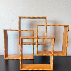 May include: A set of three interconnected light brown wooden wall shelves featuring a wavy, scalloped edge design on the outer frames. The shelves are arranged in a staggered, overlapping square configuration with smooth, polished surfaces.