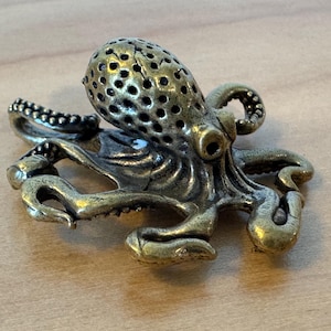 May include: A small, detailed, brass octopus figurine with a textured, patterned head. The octopus has eight tentacles and is posed in a curled position.