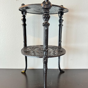May include: A black, two-tiered decorative stand with ornate detailing. The stand features a small figure at the top and intricate patterns on the circular shelves. The legs have a claw-foot design. The stand is approximately 24 inches tall.
