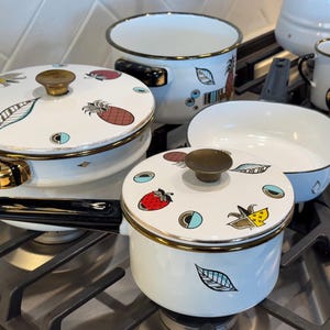 May include: A set of white enamel cookware with gold trim and colorful fruit designs. The cookware includes a saucepan, a lid, and a frying pan. The cookware is on a black stovetop.