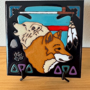 May include: Square ceramic tile with a wolf and fox design, incorporating Native American motifs. It has a black border, a turquoise background, and a red stripe. The wolf is cream, the fox is orange. Decorative triangles and a paw print are also present.