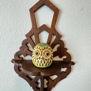 May include: A wooden wall shelf with a decorative design, holding a ceramic owl figurine. The owl is yellow and brown, with large eyes. The shelf is dark brown and has an ornate, carved design.