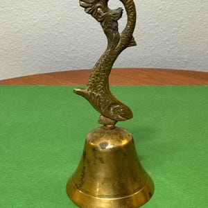 May include: A brass bell with a decorative fish-shaped handle. The bell is a traditional design and has a warm, golden color.
