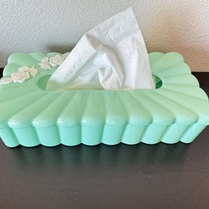 May include: A mint green tissue box with a scalloped design and white floral accents. White tissues are visible, emerging from the oval opening. The box is rectangular and sits on a dark surface.