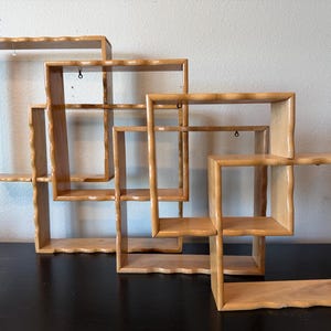 May include: A set of three interlocking square wooden wall shelves with a light brown finish. Each shelf features a distinct wavy, scalloped edge design on the outer frame and includes small metal eyelet hooks for mounting on the wall.