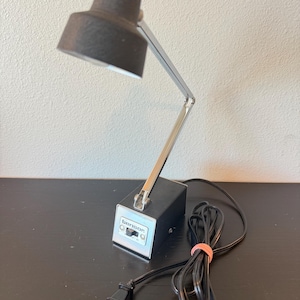 May include: A black desk lamp with a textured finish, featuring a chrome-plated adjustable arm and a rectangular base. The base displays the brand name 'benson' in white text above a black power switch. A black power cord with a plug is attached.