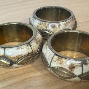 May include: Three decorative bangles with a cream-colored, patterned exterior and a gold-colored interior. The bangles have a brass-colored metal trim and decorative accents. The bangles are approximately 1 inch wide.