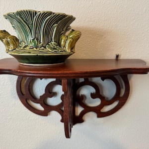 May include: A decorative, dark green ceramic planter with two frog figurines on the sides, sitting on a small, ornate, dark wood shelf. The shelf has a curved front and decorative cutouts.