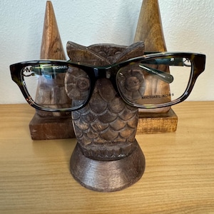 May include: A wooden owl-shaped eyeglasses holder with a pair of tortoise shell eyeglasses resting on it. The eyeglasses have the brand name "MICHAEL KORS" printed on the right temple.
