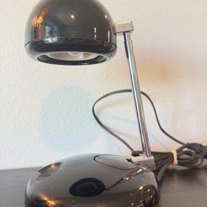 May include: A black desk lamp with a spherical head and a circular base. The lamp features a polished chrome-finished metal arm connecting the base to the light housing. A black power cord with a plug extends from the back of the lamp base.