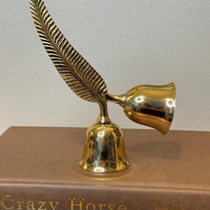 May include: A gold-colored brass bell with a feather-shaped handle. The bell is sitting on a brown book with the title "Crazy Horse The Life Behind the Legend".