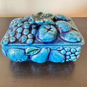 May include: A rectangular ceramic lidded box featuring a high-gloss, deep blue glaze. The lid is decorated with a three-dimensional relief of various fruits, including grapes, apples, and leaves, while the base displays additional fruit motifs.