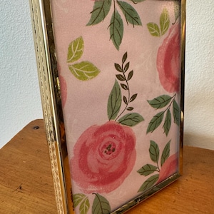 May include: A gold-toned metal picture frame with a floral print. The print features pink roses and green leaves on a light pink background. The frame is rectangular and stands upright on a wooden surface.