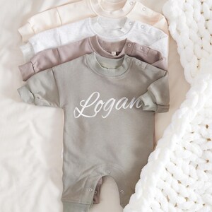 Könnte beinhalten: Ein weicher, salbeigrüner Baby-Strampler mit einem weißen gestickten Namen "Logan". Der Strampler liegt auf einem Stapel anderer Strampler in Beige, Grau und Braun.